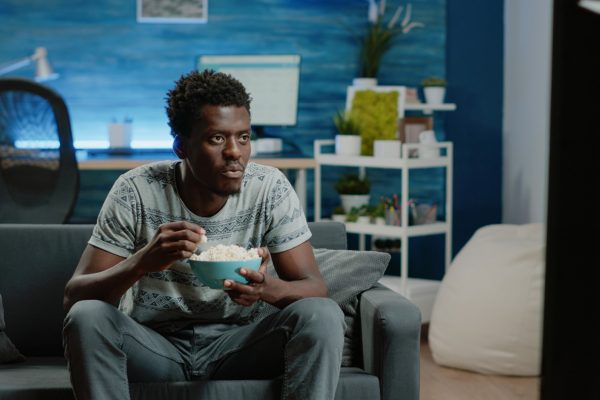 African american man enjoying comedy on television while eating popcorn at home. Black person laughing and watching movie on TV with snack in bowl. Afro adult relaxing on couch