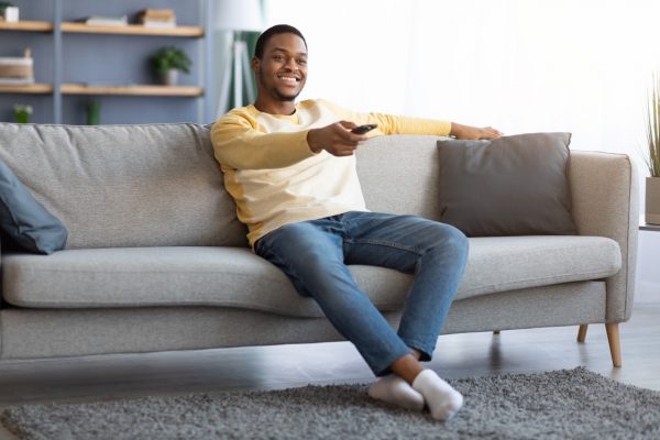 Relaxed african american guy switching channels on TV at home, holding TV remote, smiling young black man sitting on sofa in living room, watching television, enjoying lazy weekend